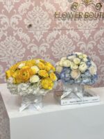 Two flower arrangements in white boxes, one yellow and one in blue and white