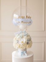 Arreglo de bienvenida al bebé: rosas marfil, hortensias y orquídea blanca en caja blanca con cinta celeste y globo burbuja