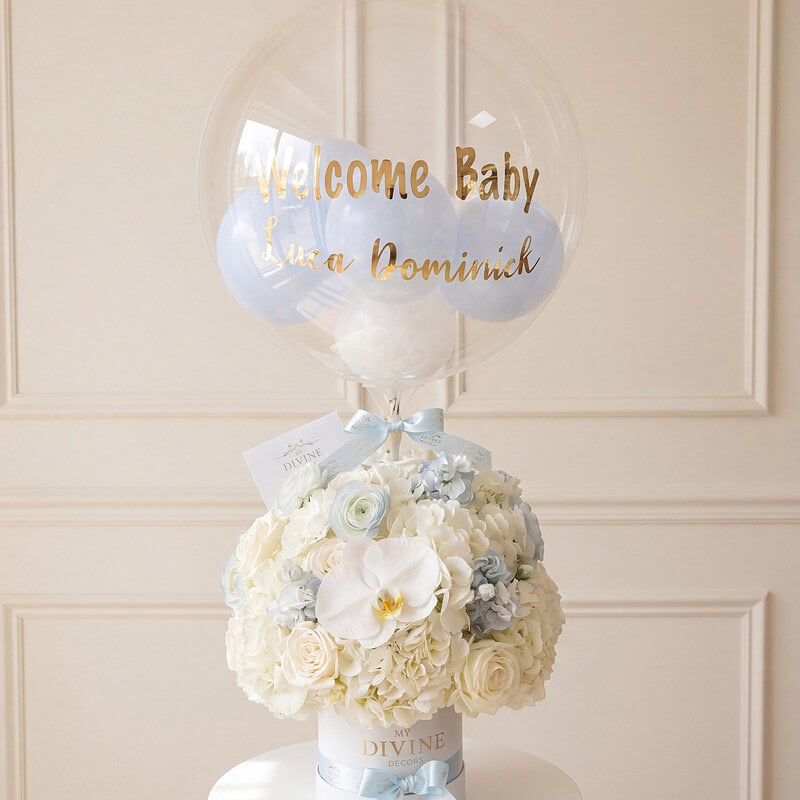 Arreglo de bienvenida al bebé: rosas marfil, hortensias y orquídea blanca en caja blanca con cinta celeste y globo burbuja