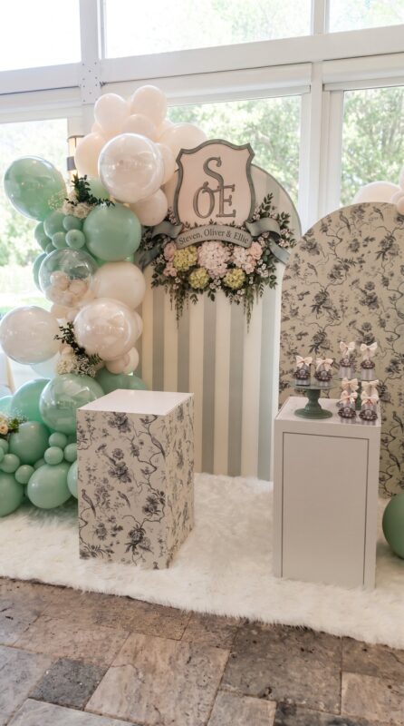 Arco de globos verde salvia y crema con escudo familiar para un bautizo en Casalina, Delray Beach