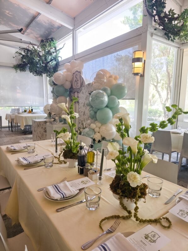 Mesa de bautizo con centros de calas y decoración con globos en verde salvia al fondo