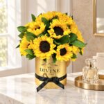 Medium Sunflower Round in a gold box with black ribbon on a marble vanity