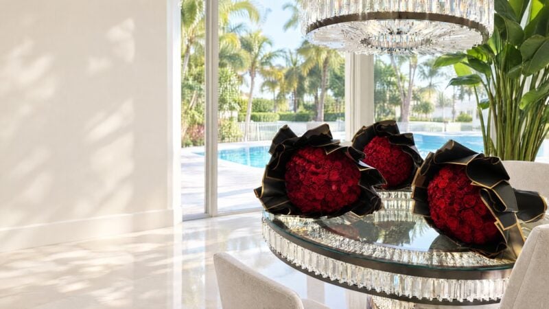 Tres ramos buchones de rosas rojas en papel negro y dorado de My Divine Decors sobre mesa de cristal, piscina y palmas al fondo