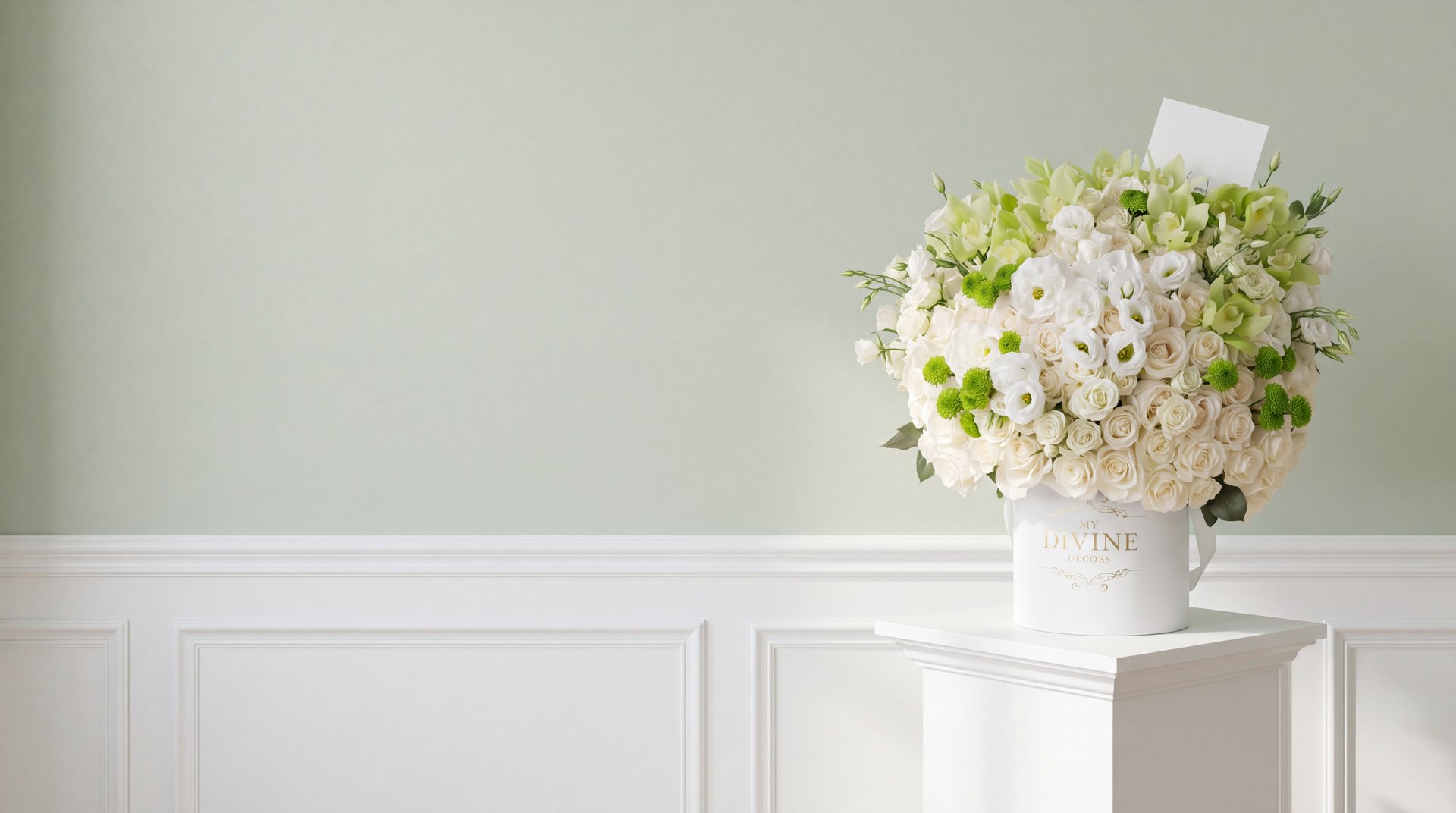 Arreglo de rosas blancas, lisianthus, orquídeas cymbidium verdes y botones verdes en una caja sombrero blanca My Divine Decors, sobre un pedestal blanco frente a una pared verde suave