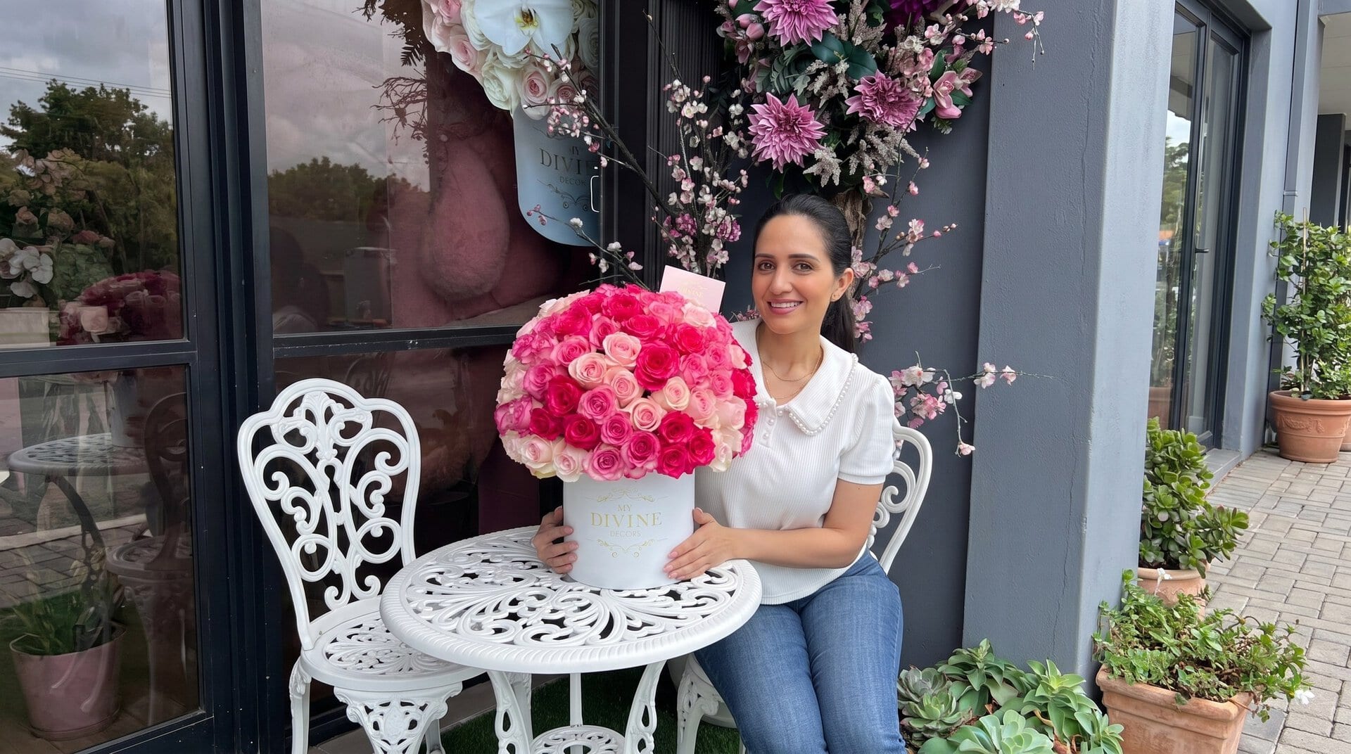 Mujer sonriendo con una caja sombrero blanca de My Divine Decors con rosas fucsia y rosadas en una terraza con flores en Miami