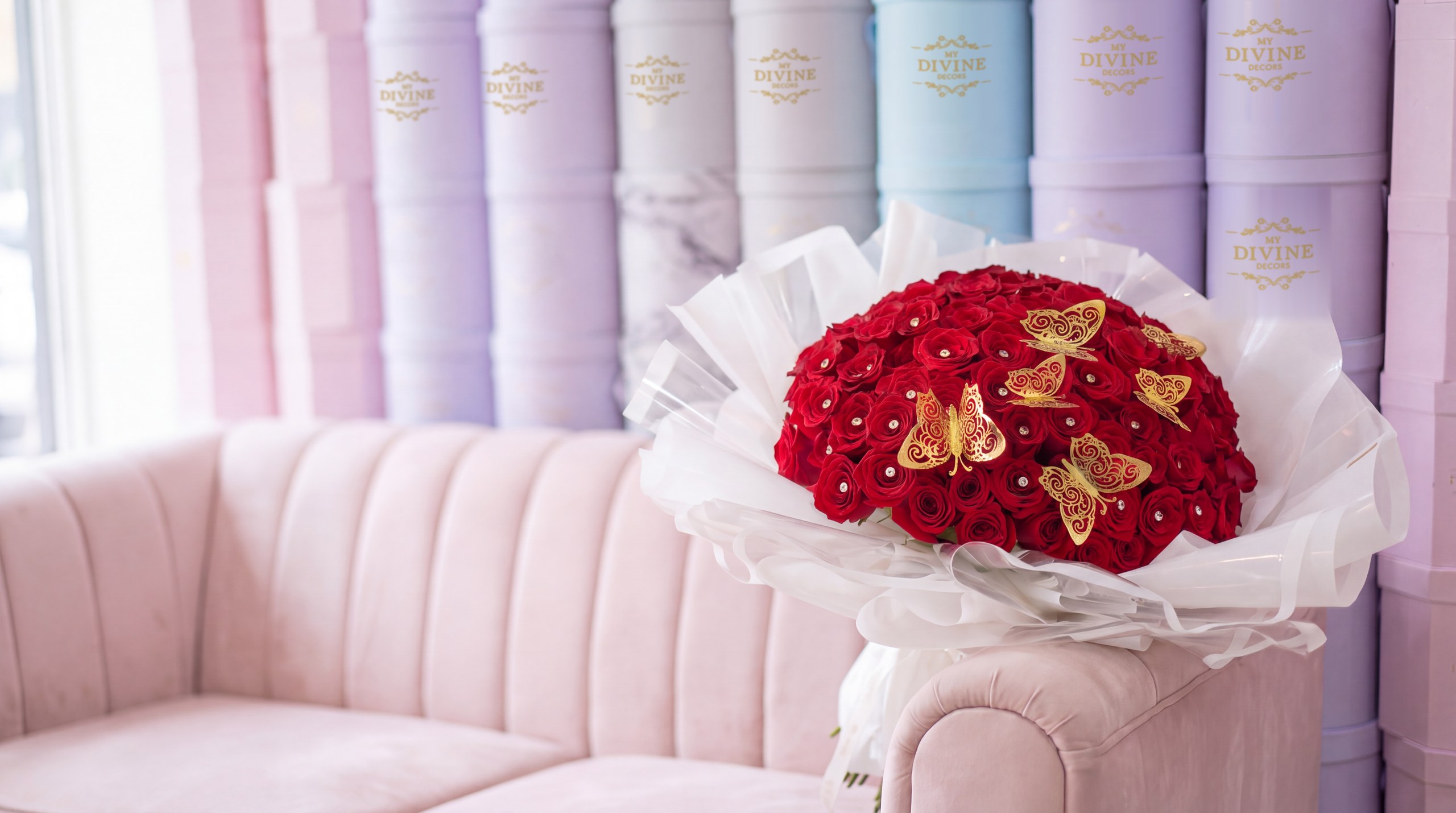 Ramo Buchon of red roses with gold butterflies by My Divine Decors on a blush velvet sofa in front of the studio wall of pastel hat boxes