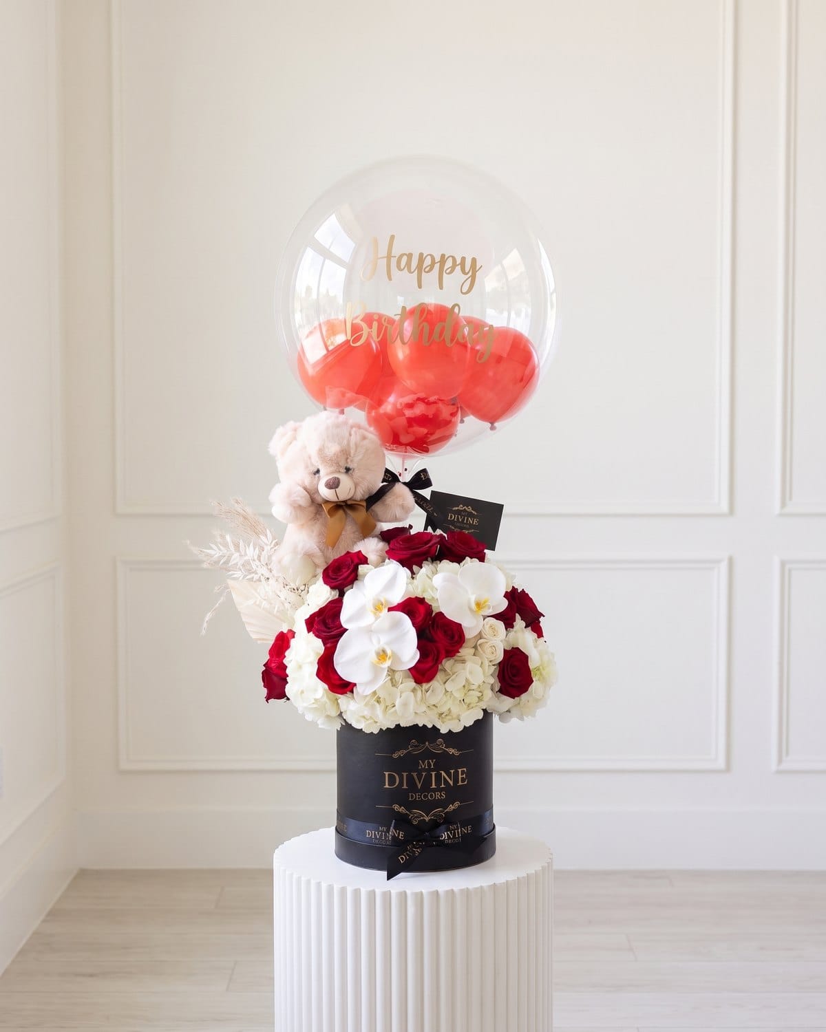 Happy Birthday bubble balloon with red balloons and a small cream teddy bear over a black MY DIVINE DECORS box of red and white roses with white orchids, on a white pedestal