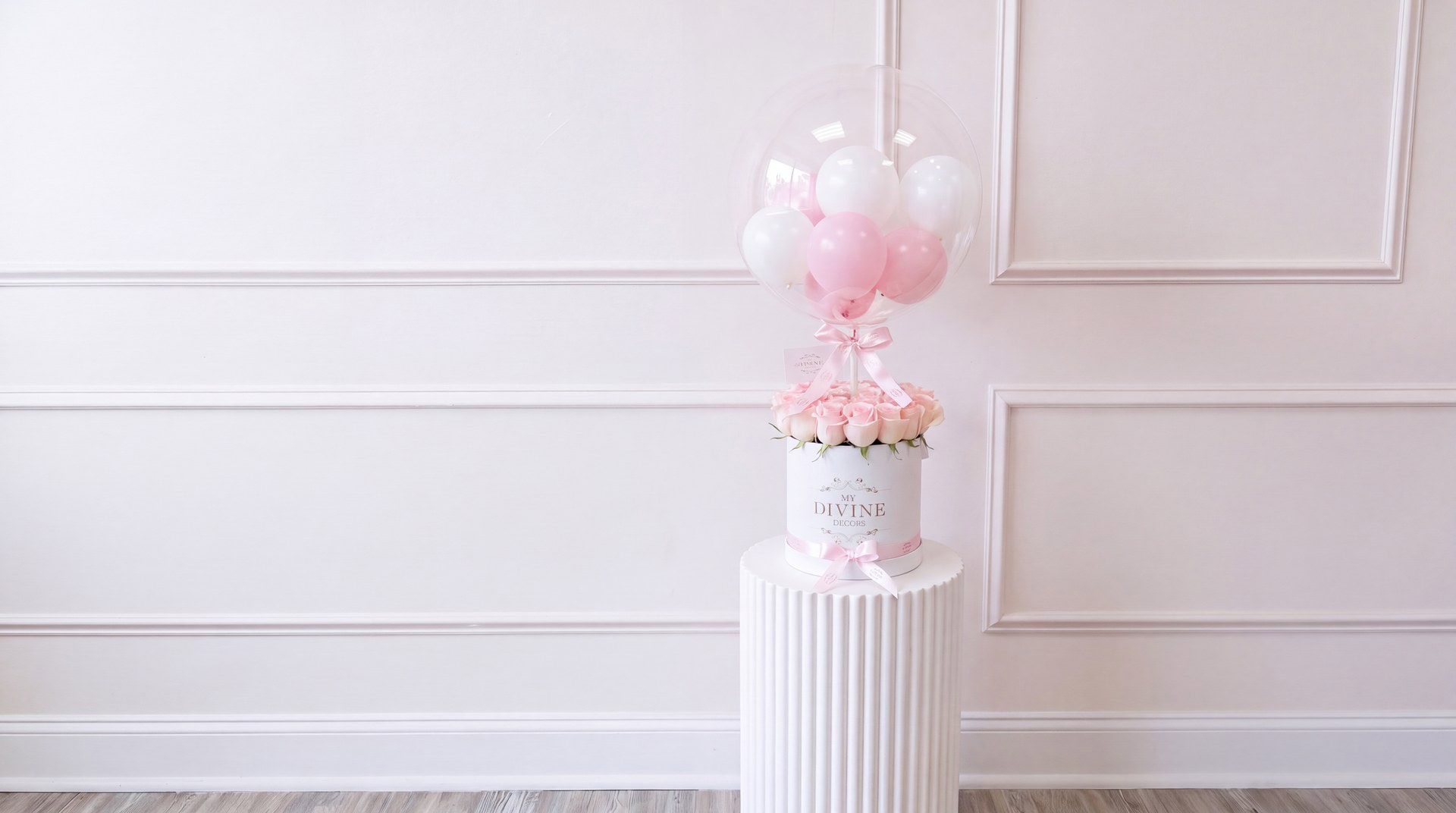 White MY DIVINE DECORS round box of pink roses under a clear bubble balloon filled with pink and white balloons, on a white pedestal in the My Divine Decors flower shop in Doral