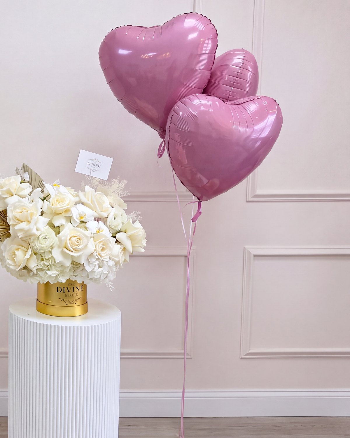 Caja dorada MY DIVINE DECORS de rosas blancas y hojas de palma secas con tres globos metálicos de corazón, sobre un pedestal blanco