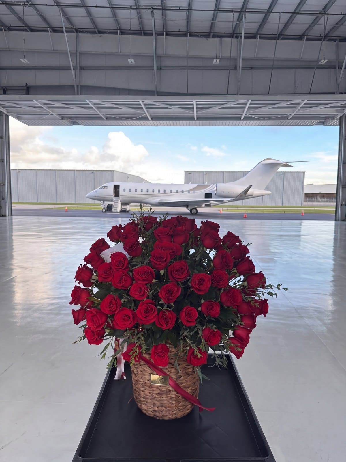 Canasta tejida de rosas rojas con cinta de satén roja sobre un carrito dentro de un hangar, con un jet privado blanco afuera, por My Divine Decors