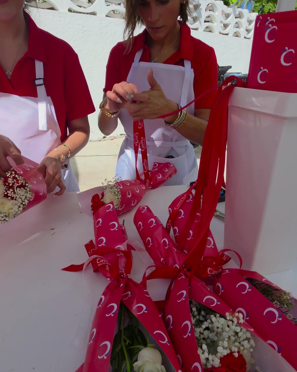 Petite bouquets of red carnations, white roses and babyโs breath in paper printed with the CarePlus logo, tied with CarePlus ribbon