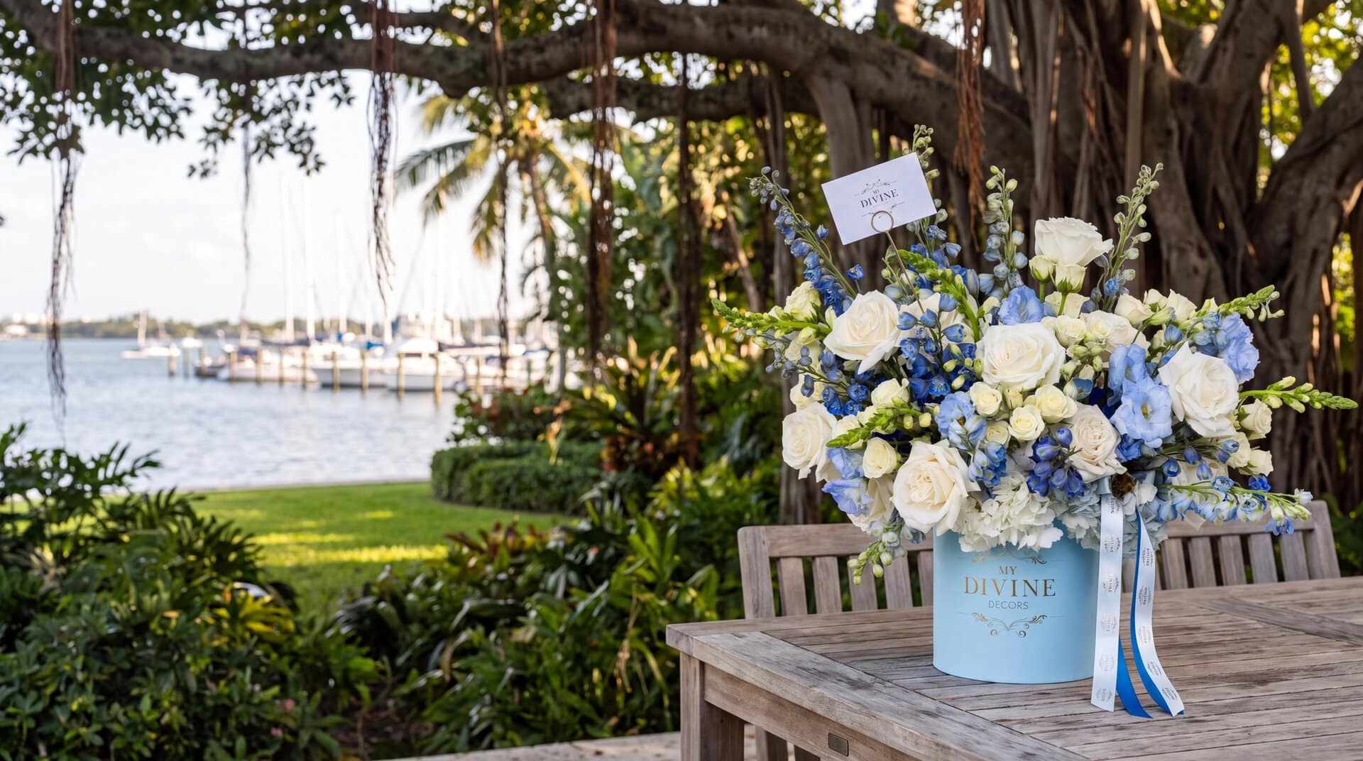 Blue Imperiale hat box by My Divine Decors with white roses, hydrangeas and blue delphinium on a teak table under a banyan tree on a Coconut Grove terrace facing the Dinner Key marina