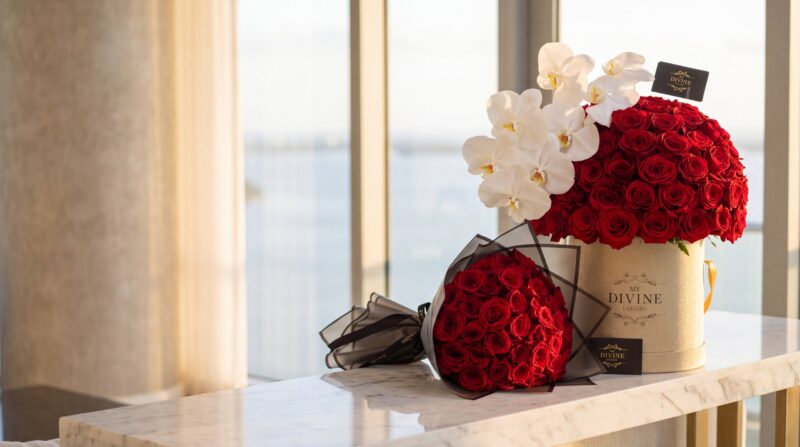 Caja signature de My Divine Decors con 100 rosas rojas y orquídeas blancas junto a un ramo buchón de rosas rojas en papel negro, sobre una consola de mármol en un penthouse de Miami