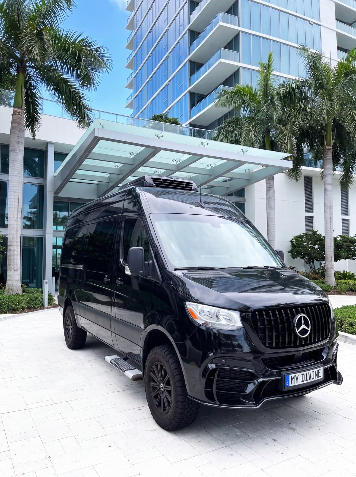 Camioneta Mercedes negra de My Divine Decors con placa MY DIVINE en la entrada de una torre frente al mar en Collins Avenue