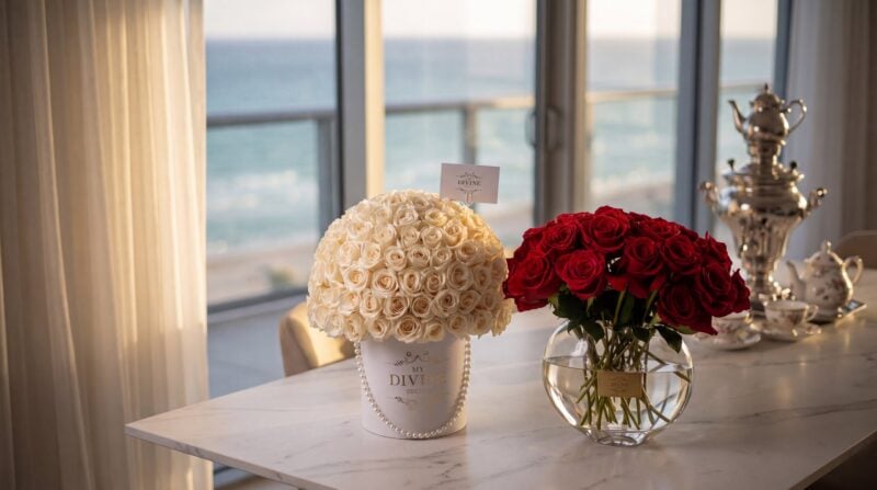 A white My Divine Decors hat box of ivory roses with a pearl string and a glass vase of long-stem red roses on a marble table in an oceanfront condo in Sunny Isles Beach, with a silver samovar and tea set behind