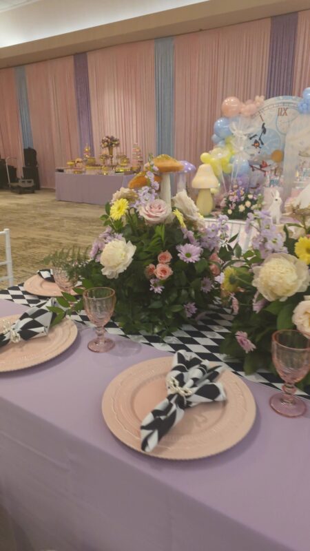 Arreglo bajo en tonos pastel con rosas y margaritas sobre una mesa lila para un primer cumpleaños de Alicia en el País de las Maravillas