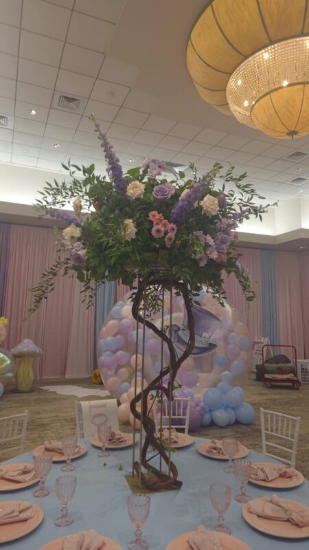 Centro alto de primer cumpleaños sobre ramas torcidas con delphinium lila y rosas rosadas en un salón de Miami