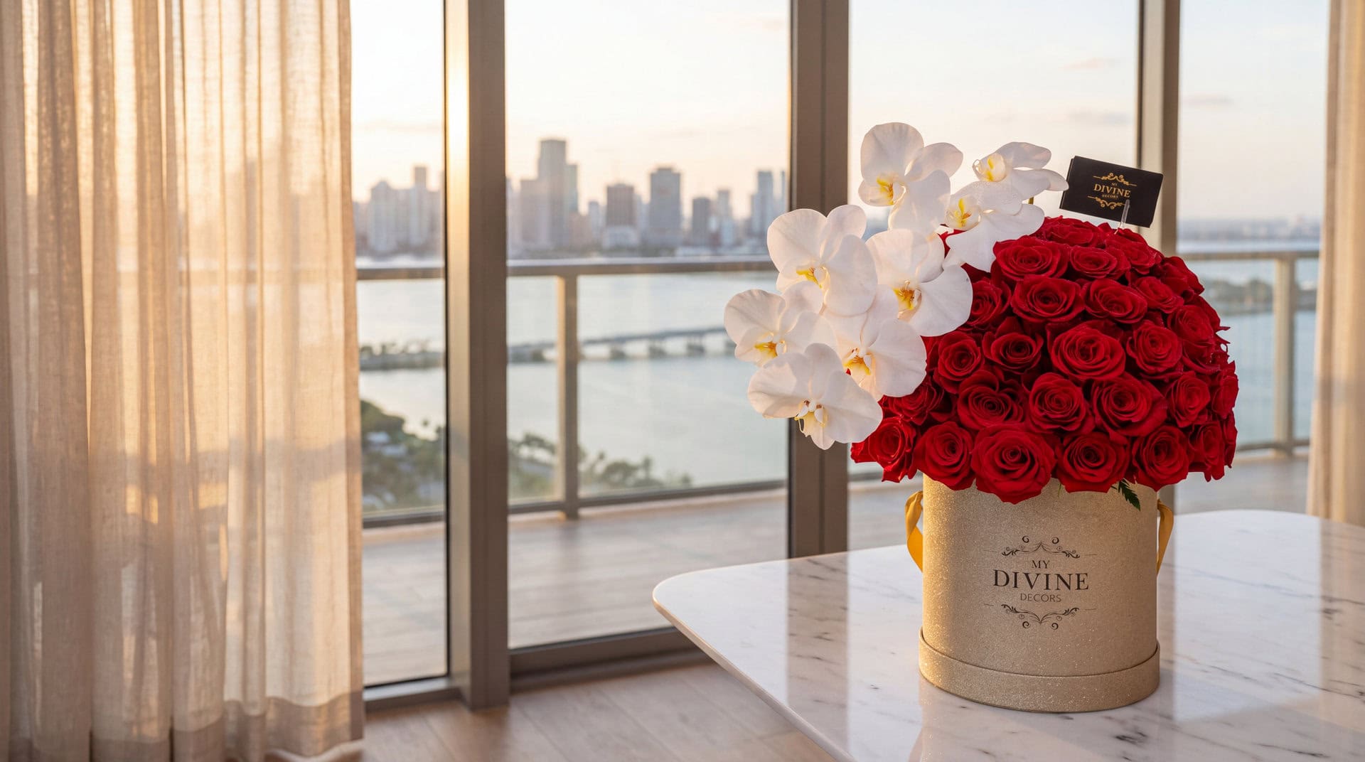 Caja dorada con cien rosas rojas y una orquídea blanca sobre una mesa de mármol en un condo de Brickell con vista a la bahía al atardecer
