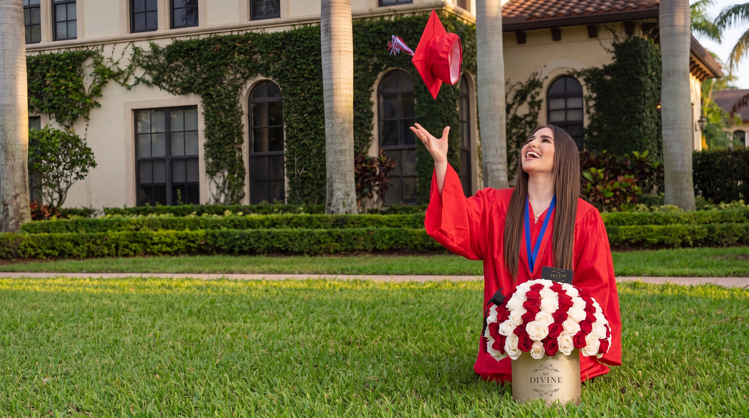 Graduada con toga roja lanzando el birrete al aire junto a una caja sombrero dorada de rosas rojas y blancas de My Divine Decors en Miami