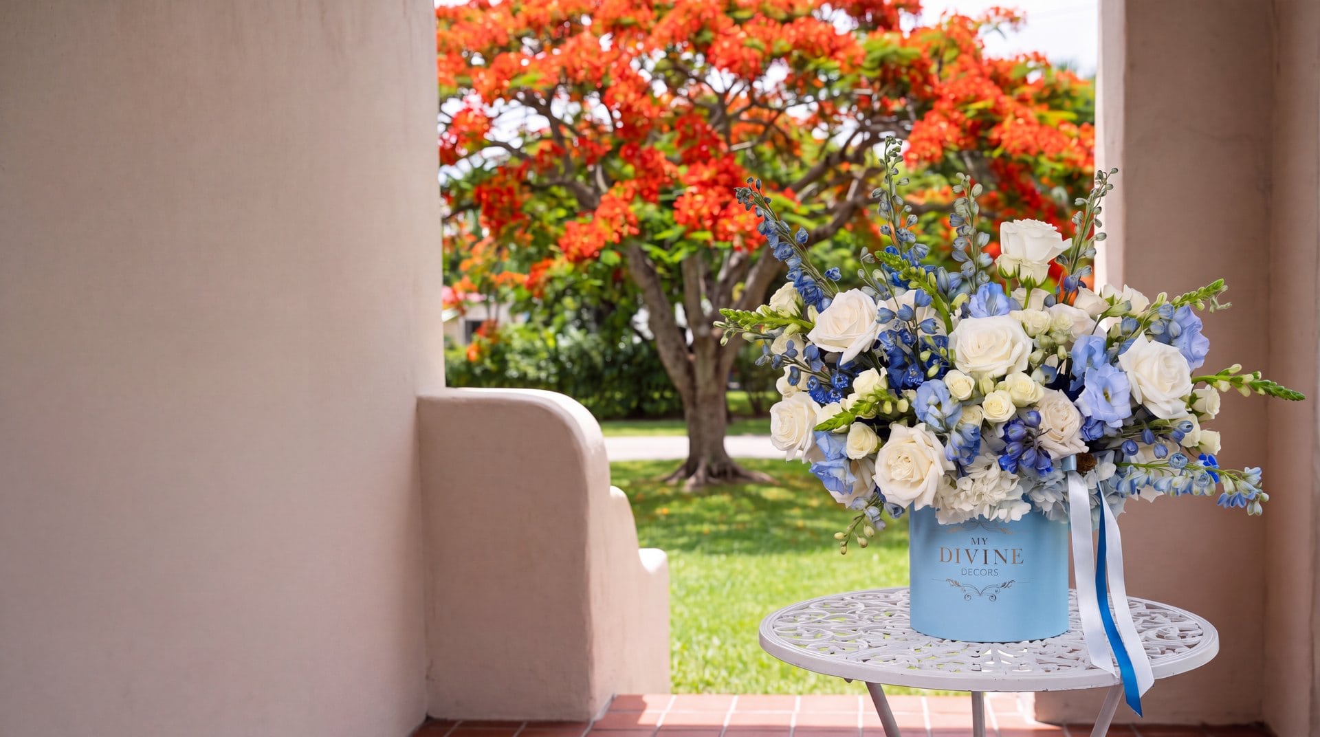 Blue Imperiale hat box of white roses, hydrangea and blue delphinium on a patio table of a Miami Springs stucco home with a royal poinciana in bloom