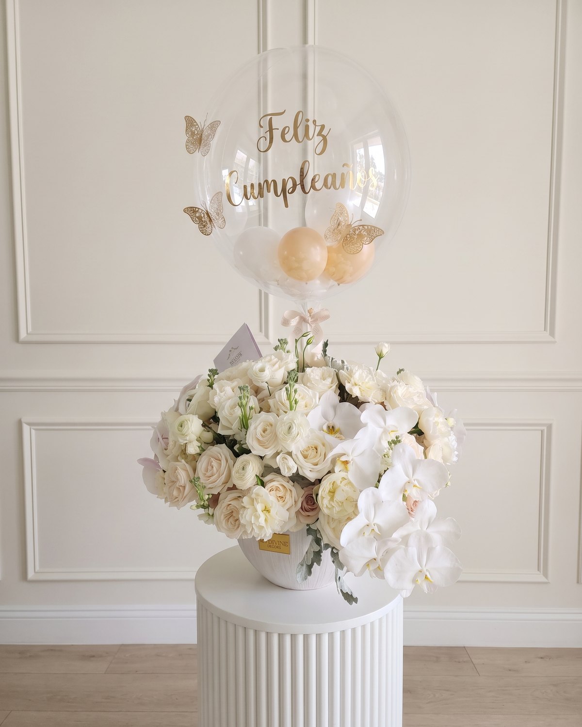 Globo burbuja de Feliz Cumpleaños con mariposas doradas sobre un arreglo de rosas blancas y orquídeas blancas, sobre un pedestal blanco