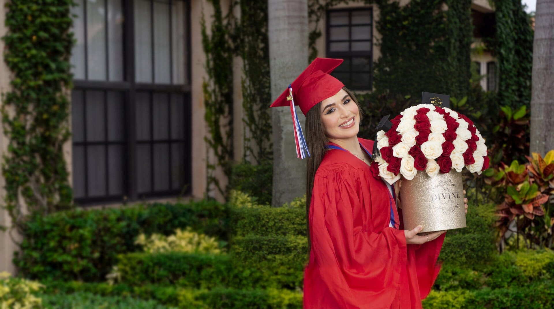 Graduate in a red cap and gown holding a gold hat box of red and white roses by My Divine Decors in a Miami garden courtyard