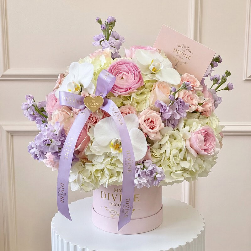 Caja Signature mediana Hanna: ranúnculos rosados, rosas blush, orquídeas blancas, hortensia y alhelí lila con cinta lila