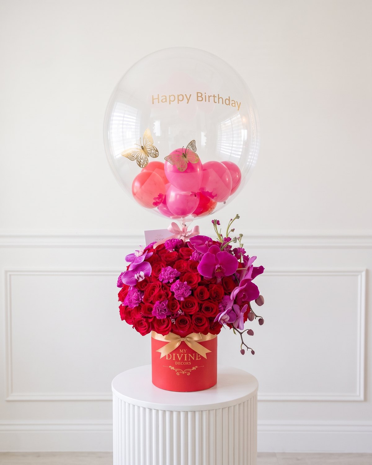 Happy Birthday bubble balloon with gold butterflies over a red MY DIVINE DECORS box of red roses, magenta orchids and fuchsia carnations, on a white pedestal