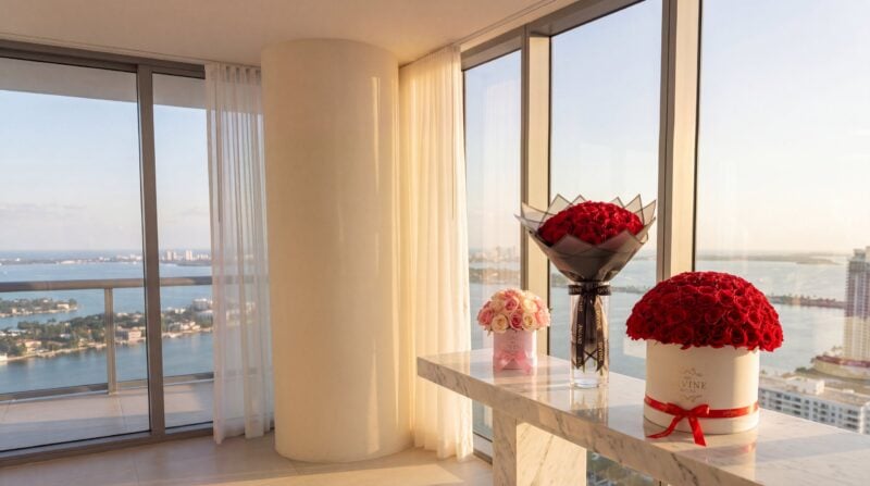 A pink My Divine Decors hat box of 25 pink and ivory roses, a red-rose Ramo Buchon in black wrap in a glass vase and a white round box of 100 red roses on a marble console in a Brickell penthouse overlooking Biscayne Bay