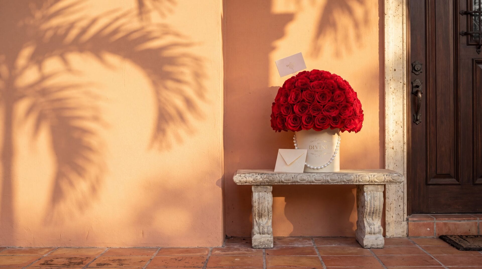 A cream My Divine Decors hat box of red roses with pearls and a card, just delivered on a stone bench by the front door of a Coral Gables home