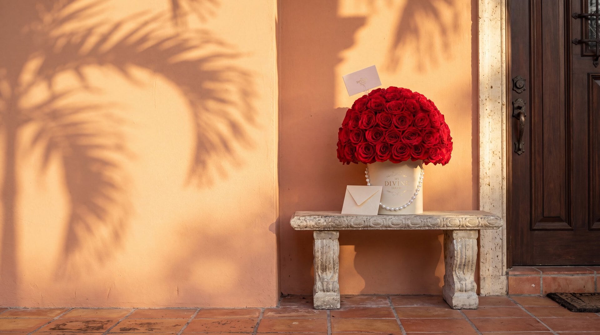 A cream My Divine Decors hat box of red roses with pearls and a card, just delivered on a stone bench by the front door of a Coral Gables home