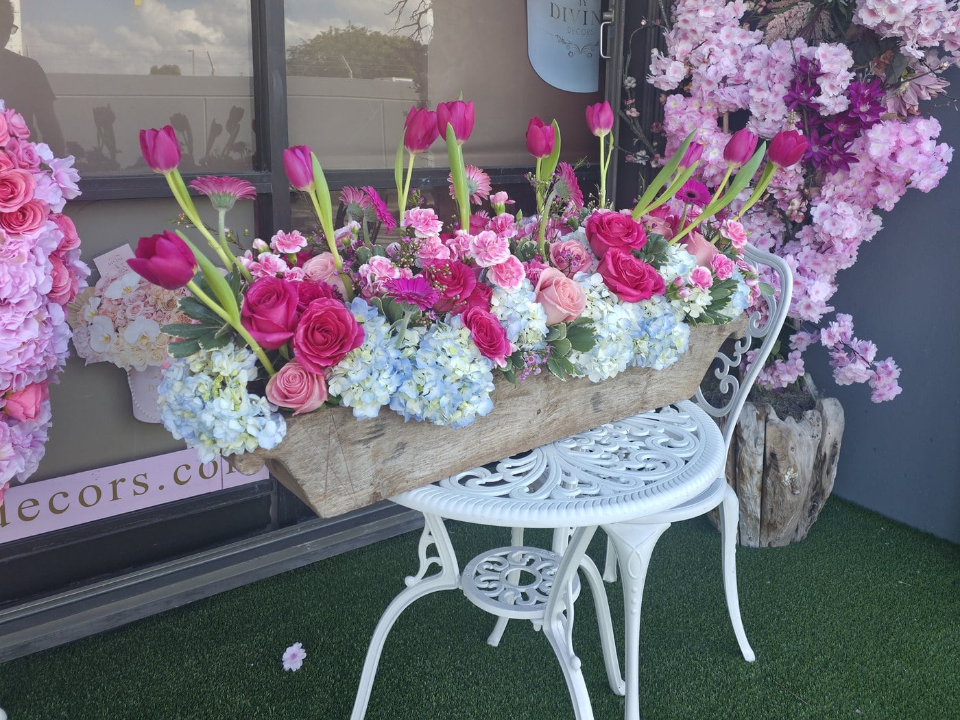 The front window of My Divine Decors in Doral with a box of pink roses, tulips and hydrangeas on a white garden table