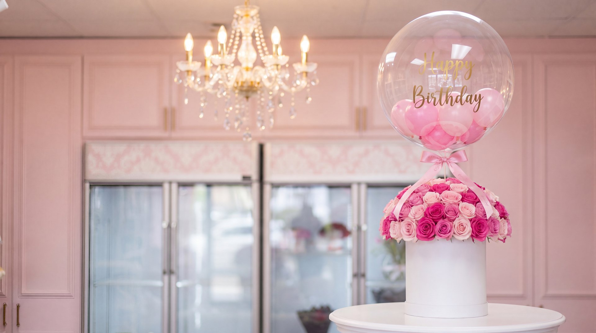 Globo burbuja transparente que dice Happy Birthday, relleno de globitos rosados, sobre una cúpula de rosas rosadas en caja sombrero blanca en nuestro taller de Doral