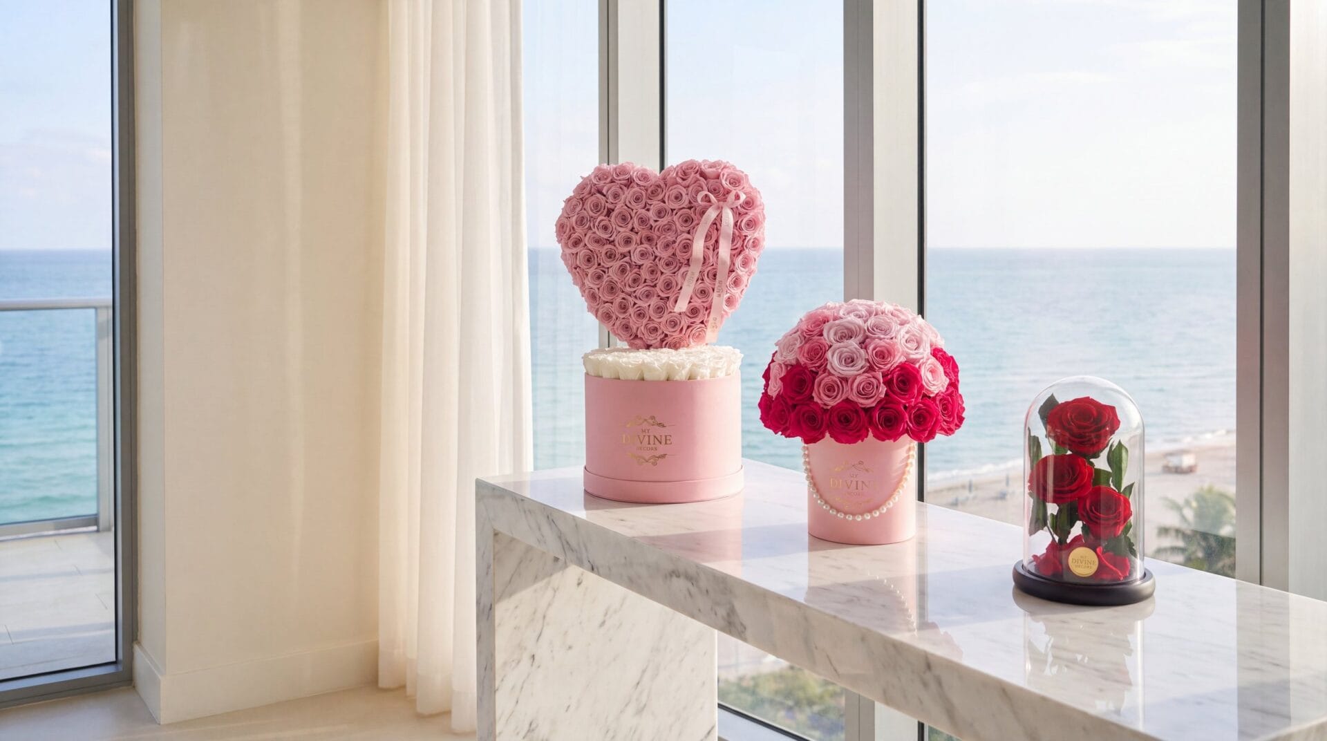 A pink My Divine Decors box with a heart of pink preserved roses, a pink hat box of pink and fuchsia preserved roses with a pearl string and a glass dome of three red preserved roses on a marble console in a Bal Harbour condo overlooking the ocean