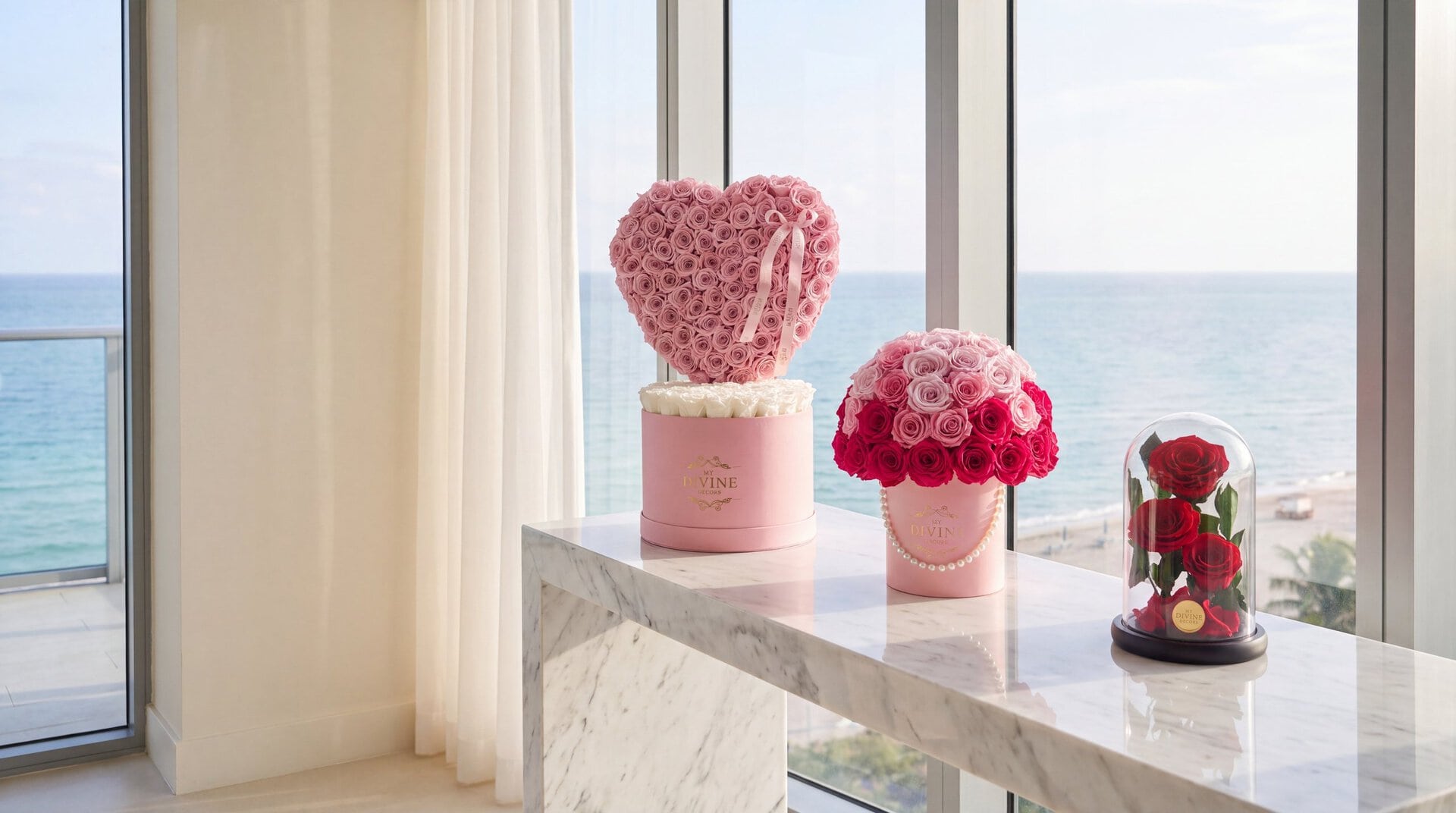 A pink My Divine Decors box with a heart of pink preserved roses, a pink hat box of pink and fuchsia preserved roses with a pearl string and a glass dome of three red preserved roses on a marble console in a Bal Harbour condo overlooking the ocean