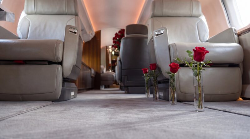 Single red roses in clear bud vases standing on the aisle of a private jet cabin, with a large red rose arrangement at the far end, by My Divine Decors in Miami