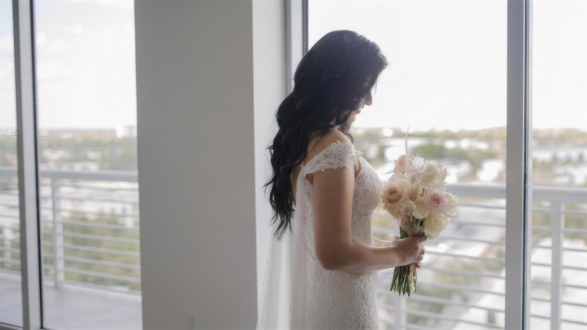 Novia con vestido de encaje junto a la ventana de un piso alto en Miami con un ramo de novia de rosas blancas y rosadas y pampas de My Divine Decors