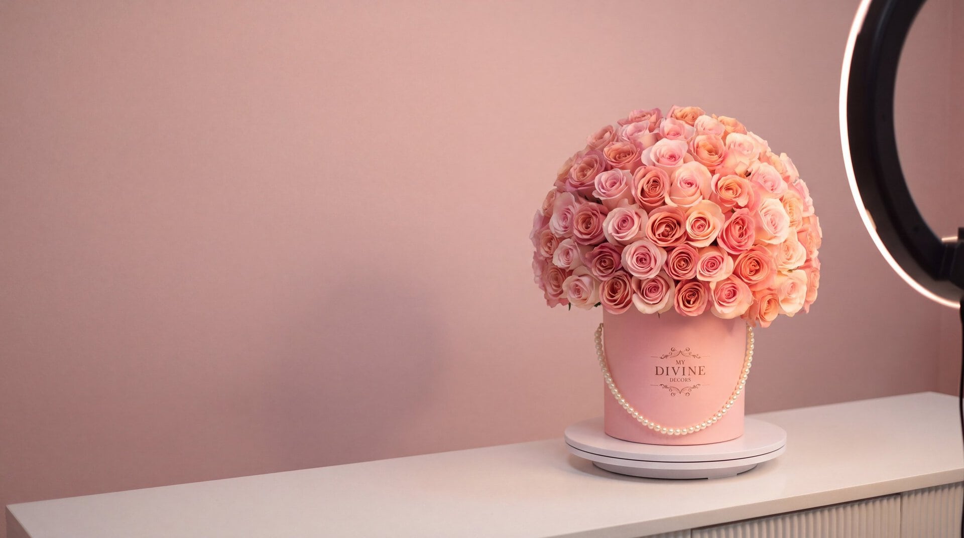 Una caja rosada de My Divine Decors con rosas rosadas y melocotón y perlas sobre un plato giratorio bajo un aro de luz, lista para grabarse en nuestro taller de Doral