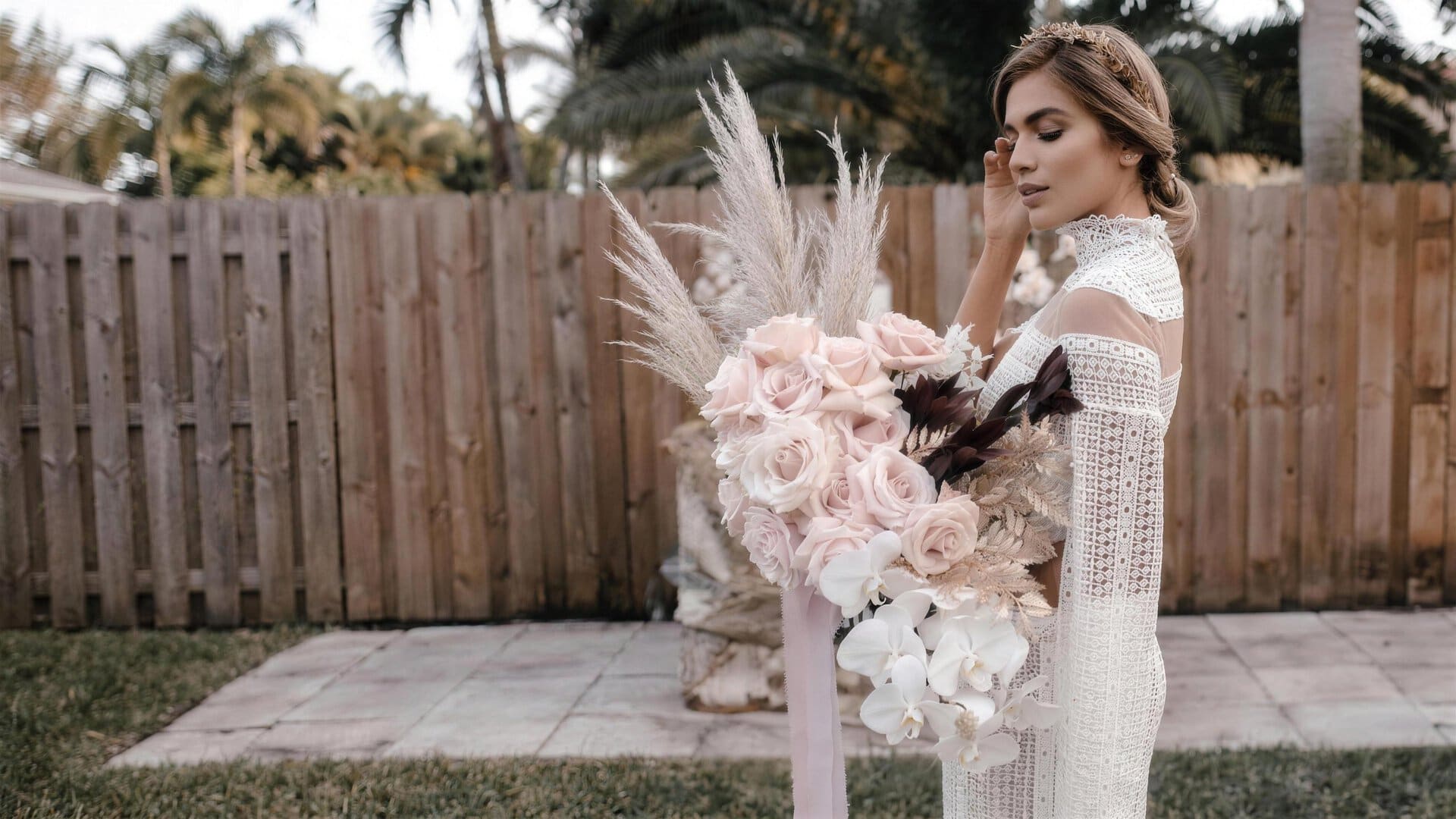 Bride in a white lace gown holding a cascading bridal bouquet of blush pink roses, white orchids and pampas grass by My Divine Decors in a Miami garden